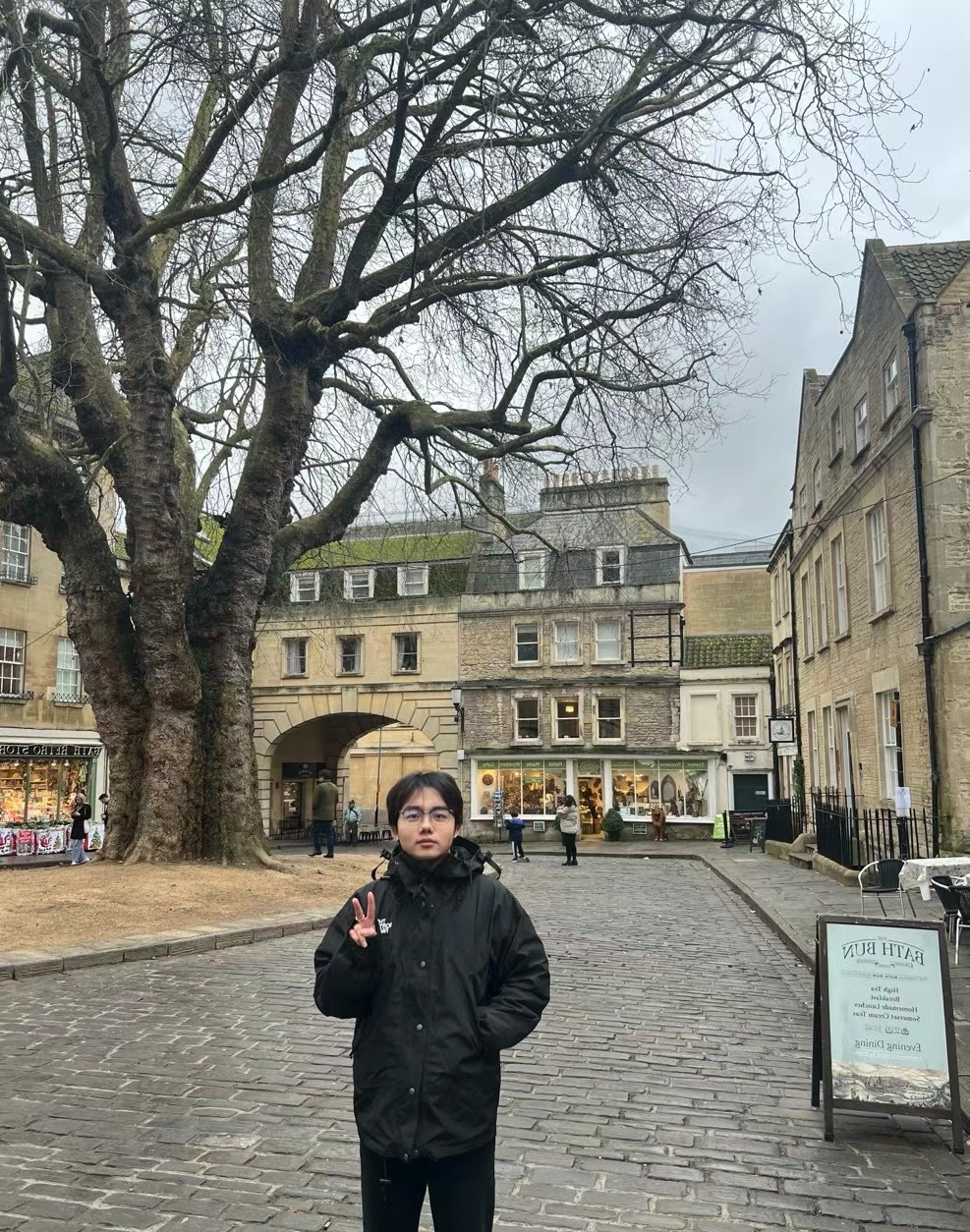 Yipeng Chen standing in a cobbled square beside a large tree and historic stone buildings.